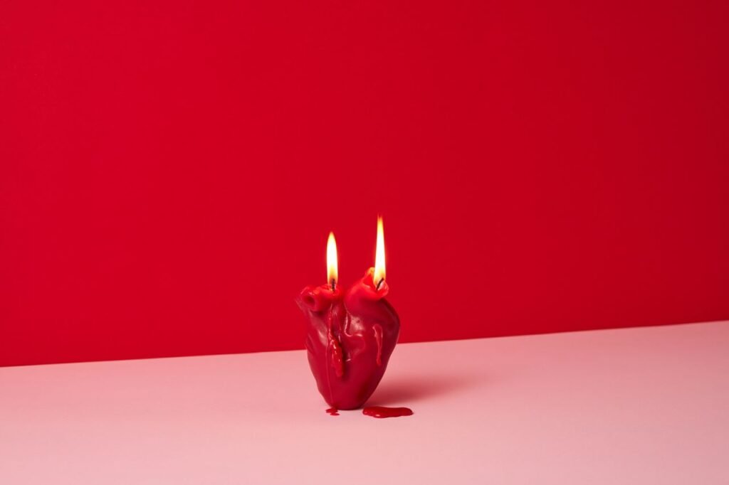 Heart-shaped red candle with dual flames on a pink surface against a red background, evoking romantic ambiance.